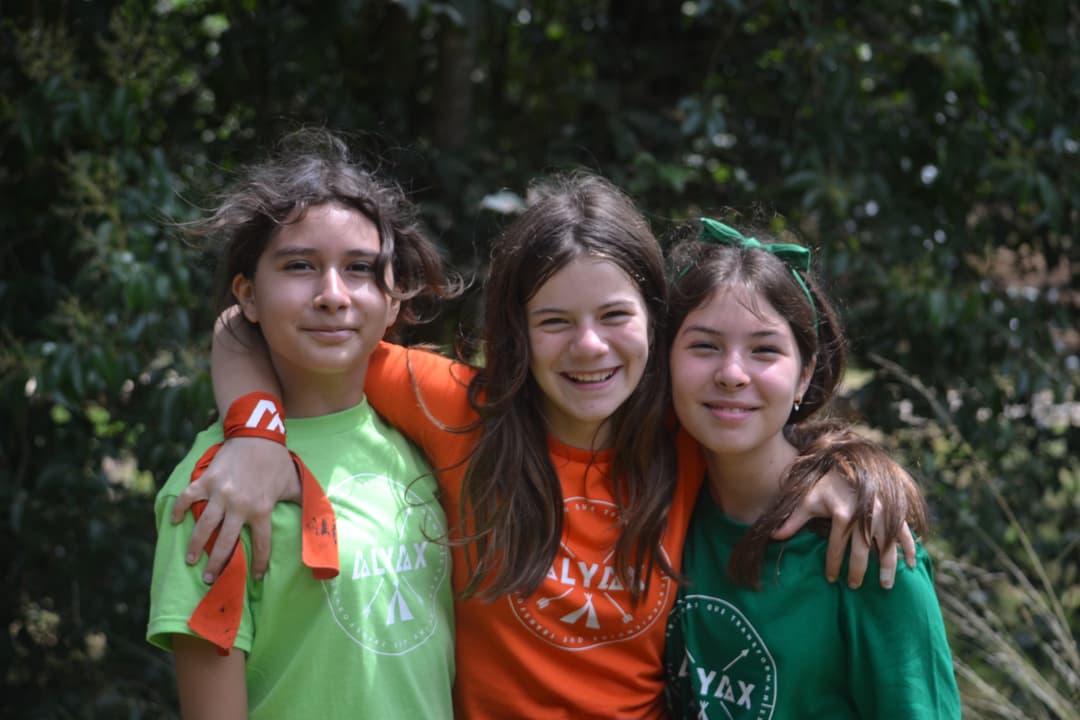 Tres niños sonrientes con pintura facial y collares de colores, destacando la amistad y el espíritu festivo del campamento
